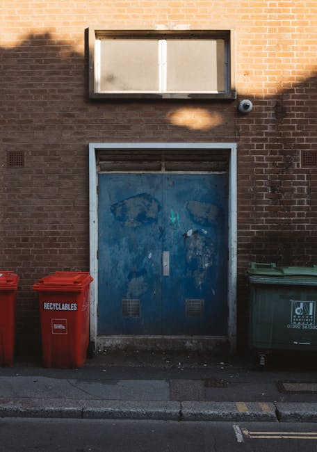 A weathered blue metal door set into a brick wall with visible signs of wear, including peeling paint and patches of rust. Above the door, there is a rectangular window with frosted or dirty glass, framed by dark metal, allowing minimal light to pass through. To the right of the door, a small black ventilation vent protrudes from the brickwork. On the pavement directly in front of the door, there are three coloured waste bins: a red recycling bin on the left, with a clear label indicating recyclables, and a larger green bin to the right, both made of plastic with smooth surfaces and fitted with lids. The red bin appears slightly more prominent due to its placement, while the green bin has a partially visible logo or text. The scene is illuminated by natural daylight, casting soft shadows on the brick wall and pavement, creating a straightforward but realistic view of an urban external service point for waste collection, potentially used for private rubbish disposal or on-site clearance activities, as supported by the presence of the labelled waste containers nearby.