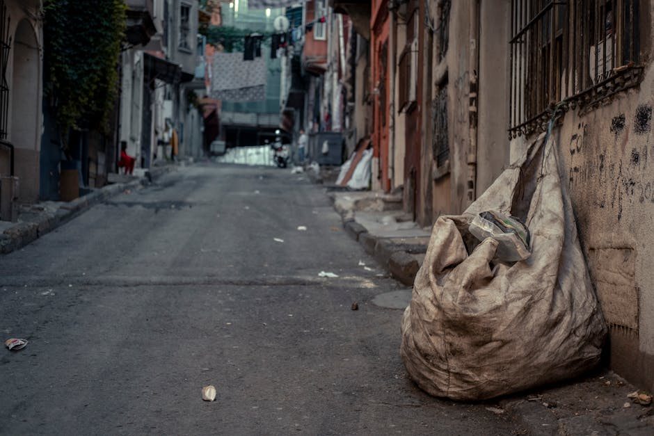 A large, dirty, beige cloth bag filled with household waste and litter, situated on a narrow urban street pavement. The bag appears worn and stained, with some plastic waste visibly protruding from the top. The street is lined with aging residential buildings featuring metal bars on windows and graffiti on the walls, indicating a densely populated area. In the background, washing lines with hanging laundry stretch between buildings, and the street is slightly inclined, with uneven asphalt and small scattered debris along the edges. The scene is illuminated by natural light, casting soft shadows on the ground, and conveys a context where independent rubbish collection might be necessary for waste disposal in tight or inaccessible urban spaces. The overall setting reflects a typical local environment requiring efficient waste removal services, such as those offered by wastecollectionkingstonuponthames.co.uk, to manage rubbish in hard-to-reach streets and courtyards.