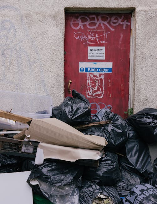 A pile of black rubbish bags and discarded materials, including cardboard and plastic items, stacked against a beige rough-textured external wall beneath a red metal door covered in graffiti. The door has a small sign that reads 'NO MONEY' and a larger blue and white sign stating 'Keep clear.' The scene suggests an area of informal waste disposal or an overflow of waste, with some waste spilling onto the ground and partially blocking the lower part of the door. The environment appears to be an urban street or alleyway, with the waste occupying a small section of the pavement or entrance, and the surrounding surfaces showing signs of wear and weathering. This image exemplifies a situation where external rubbish accumulation may require professional waste removal services, such as those offered by Kingston upon Thames waste management providers like wastecollectionkingstonuponthames.co.uk, focusing on private or alternative clearance options in urban environments.