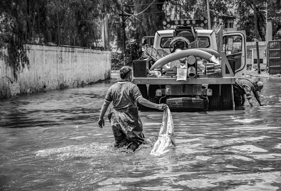 In the foreground, a man wearing waterproof overalls stands waist-deep in floodwater, holding a plastic bag used for collecting debris. Behind him, another individual also clad in waterproof clothing is partially submerged and appears to be working alongside a large utility vehicle. The vehicle is parked on the flooded street, with its rear section loaded with equipment, hoses, and containers associated with flood recovery and waste removal. The scene is set in an urban area, with a concrete wall on the left and a metal gate on the right, both partially submerged or surrounded by water. The environment suggests a recent flood event requiring emergency waste management or flood debris clearance, with the water's surface rippling from the activity. The black-and-white photograph emphasizes the textures of the water, clothing, and vehicle, illustrating the efforts involved in flood recovery and potential private waste disposal services, such as those provided by companies like wastecollectionkingstonuponthames.co.uk, which often assist with alternative waste handling after emergencies or flooding.