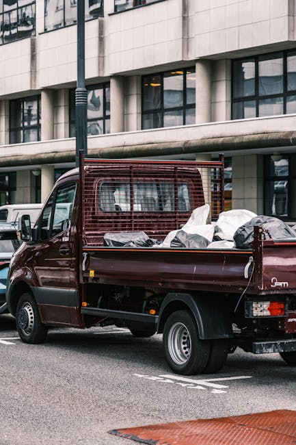 A small burgundy flatbed truck parked on an urban street in front of a modern office building with large glass windows and a white exterior. The truck's cargo area is filled with various types of rubbish, including black and white plastic bags, some of which are partially open revealing waste inside. A metal protective grille is mounted behind the driver’s cab, providing security for the cargo, and the truck features a compact, utilitarian design with a black bumper, silver wheel rims, and clear tail lights. The surrounding environment includes a streetlamp nearby and other vehicles parked further along the road. The scene reflects an instance of private waste collection or rubbish removal service, consistent with services offered by companies like wastecollectionkingstonuponthames.co.uk, emphasizing alternative waste handling methods in urban settings while maintaining a neutral, functional tone aimed at supporting accessibility and SEO relevance in rubbish disposal contexts.