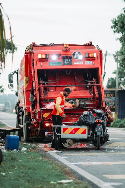 A worker in a high-visibility yellow and orange vest is seen operating a large red waste collection truck equipped with hydraulic arms and loading mechanisms, positioned on the roadside. The truck has reflective striping and identification labels, with a partially open rear compartment revealing collected rubbish. The worker is handling a black plastic bag and appears to be loading or managing waste debris, which is scattered on the ground nearby. The scene is set on a paved street with a narrow strip of grass and a sidewalk in the foreground, and there are trees and utility poles visible in the background, suggesting an urban or suburban environment. The weather appears overcast, with diffuse lighting contributing to a neutral, professional atmosphere associated with waste management operations, reflecting the work of companies such as waste collection services in Kingston or similar settings focused on private or independent rubbish removal and on-site clearance tasks.