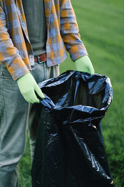 A person dressed in a casual plaid shirt, green gloves, and beige pants is holding open a large black plastic rubbish bag outdoors on a grassy area. The person's left hand is gripping the top edge of the bag, which is partially folded over, revealing its shiny, smooth plastic interior. The background shows a well-maintained green lawn, suggesting a garden or park setting. The scene appears to depict an activity related to waste collection or rubbish removal, possibly as part of a private clean-up task or eco-friendly initiative. The overall environment is bright and natural, with no other objects or people visible in the image. This visual context aligns with independent waste handling or alternative rubbish disposal methods, which companies like wastecollectionkingstonuponthames.co.uk may support for residential or small commercial projects in Kingston upon Thames.