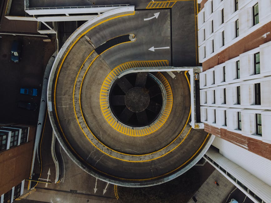 An aerial view of a multi-level, circular concrete car park with yellow safety markings along the edges and on the central ramp. The ramp curves around a central void, which appears open or shaded, with a dark, textured surface. Surrounding the structure are adjacent buildings with modern white and brick facades, and a few parked vehicles are visible in the lower left corner. The parking area features clear directional arrows painted on the surface, guiding traffic flow. The scene is evenly lit with natural daylight, highlighting the textures of the concrete and the sharp lines of the painted markings. This overhead view showcases the compact design of the parking structure, which could be part of an urban environment requiring efficient space use, aligning with services like private waste management, including exteriors and access areas that may need regular rubbish clearance. The image emphasizes the architectural and functional aspects of urban infrastructure, relevant to efficient logistics and waste management in busy city environments, potentially involving independent collection or site clearance processes through waste collection services like those offered by wastecollectionkingstonuponthames.co.uk.
