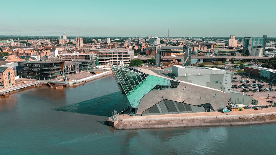 An aerial view of a modern, angular building situated on the waterfront, featuring a distinctive sloped glass roof with a greenish tint, metal cladding in shades of gray, and a concrete base along the water's edge. The building is surrounded by a paved area with some parked vehicles and a small dock extending into the water. In the background, there are various cityscape elements, including residential and commercial buildings, some with pitched roofs and others with flat roofs, along with cranes and construction activity. The overall scene is illuminated by daylight, highlighting the contemporary architectural design of the structure and the urban environment, which relates to waste collection or management services such as those provided by [COMPANY_NAME] in the context of private waste disposal or on-site clearance near urban development areas.