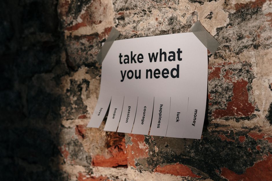 A white paper sign with bold black text reading 'take what you need' is taped to a weathered exposed brick wall. The sign features a list of options including 'hope,' 'passion,' 'courage,' 'happiness,' 'luck,' and 'money,' each with a small checkbox underneath. The paper appears slightly curled at the edges and is affixed with grey adhesive tape at the top corners. The background consists of rough, uneven brickwork in shades of red, grey, and black, with some mortar and chipped areas visible, suggesting an aged or industrial environment. The overall composition suggests an inviting, community-based message in a space that could relate to shared or private waste and resource sharing, subtly aligning with themes of alternative disposal or on-site clearing potentially managed by waste collection services from Kingston upon Thames. The scene is illuminated with natural or soft artificial light, emphasizing the textures and muted tones of the brick surface and the contrast of the pristine paper sign.